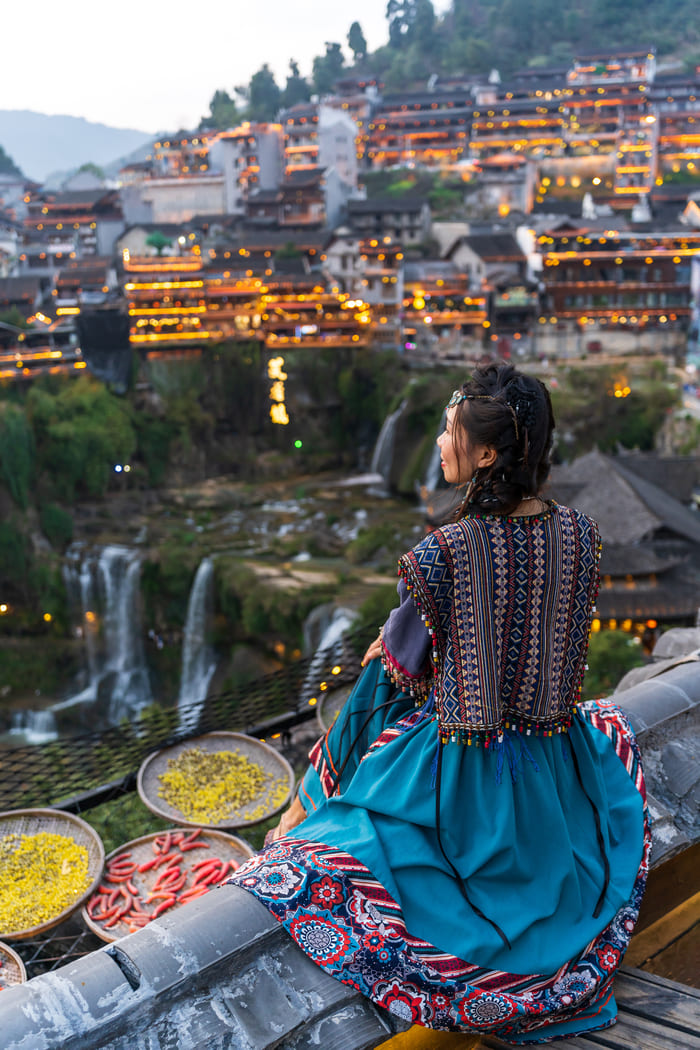 Mujer con traje tipico color azul dando la espalda, sentada viendo la ciudad de Lijiang
